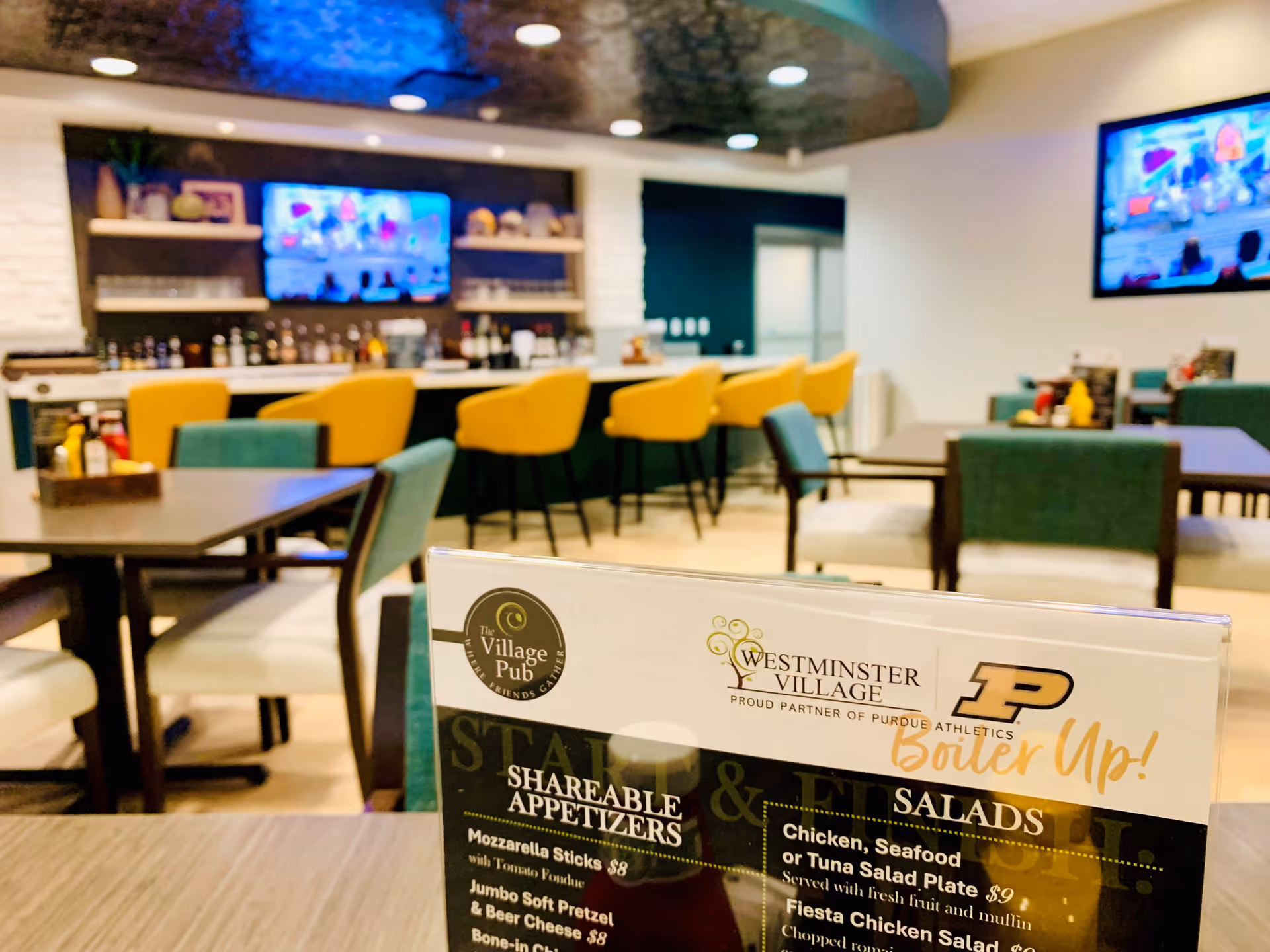 Interior view of a dining area in Westminster Village featuring tables and chairs with a menu card in the foreground. The background shows a bar with yellow bar stools and a television mounted on the wall.