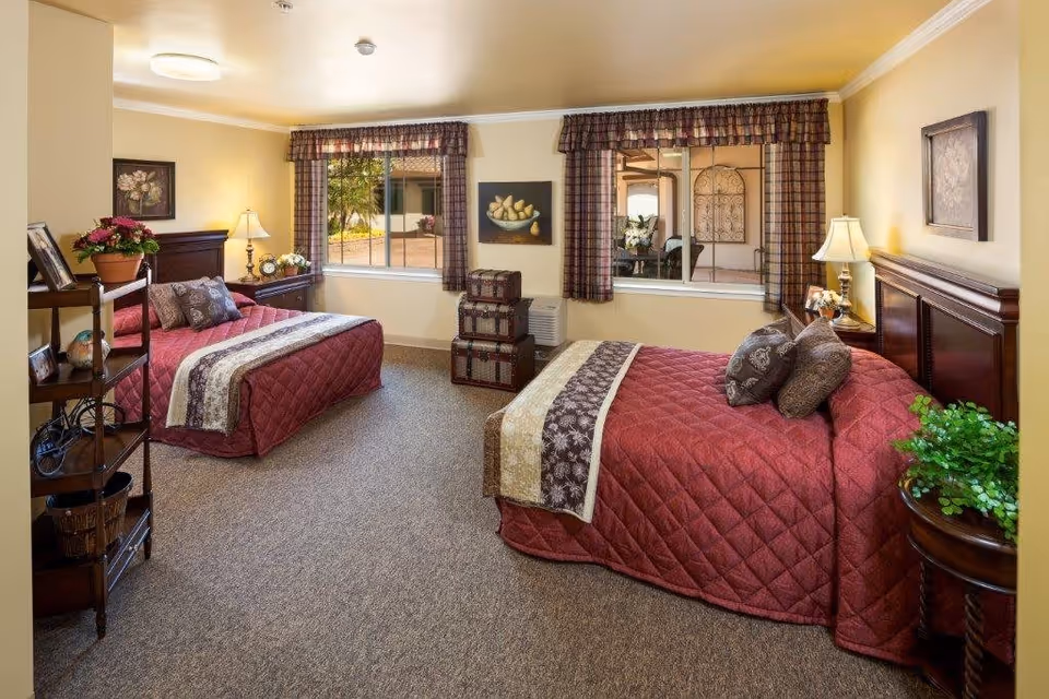 A cozy bedroom with two beds covered in red quilted bedspreads and decorative pillows. The room has beige walls, carpeted floor, two windows with plaid curtains, and wooden furniture including nightstands with lamps, a small shelving unit, and decorative plants and artwork on the walls.