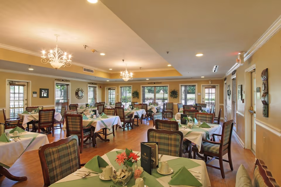 Well-lit dining room with multiple tables set with white tablecloths and green napkins, chandeliers, and upholstered chairs.
