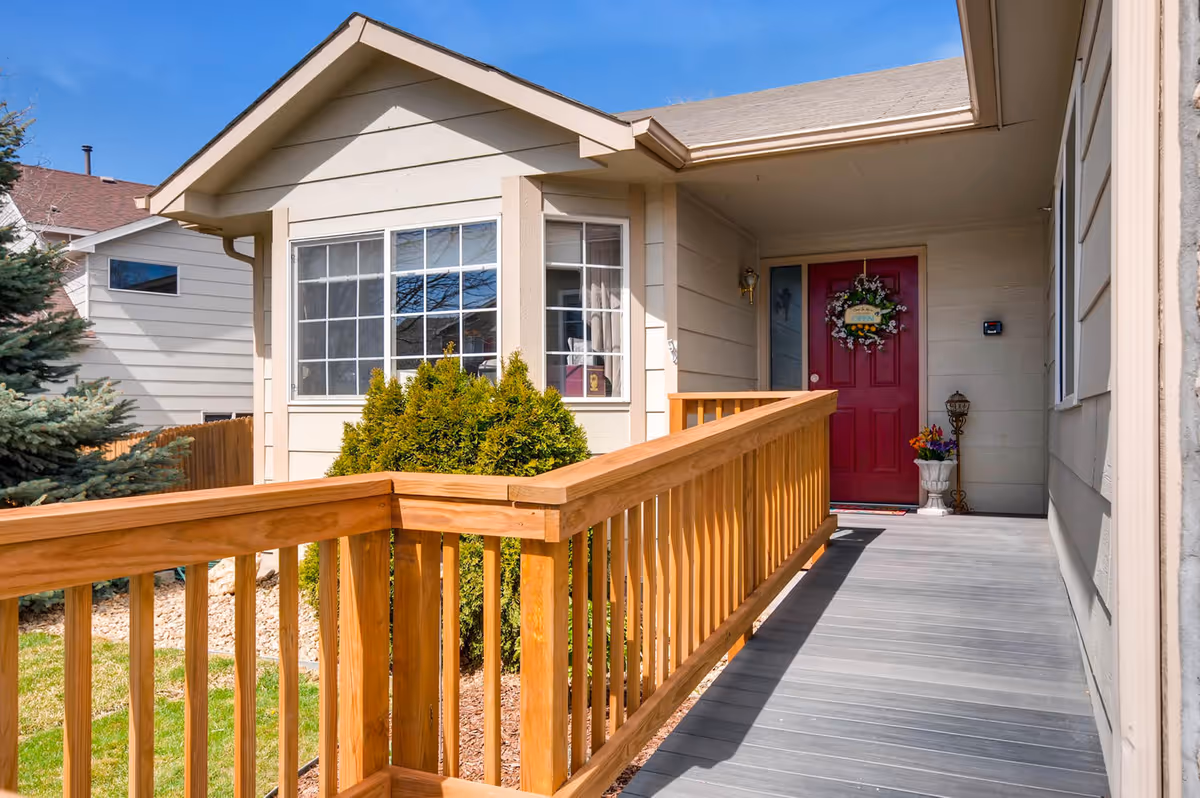 Front entrance of a single-story house with a wooden ramp leading to a red door decorated with a floral wreath. There are shrubs and a small garden area beside the ramp, and the exterior walls are light beige with white trim. The sky is clear and blue.