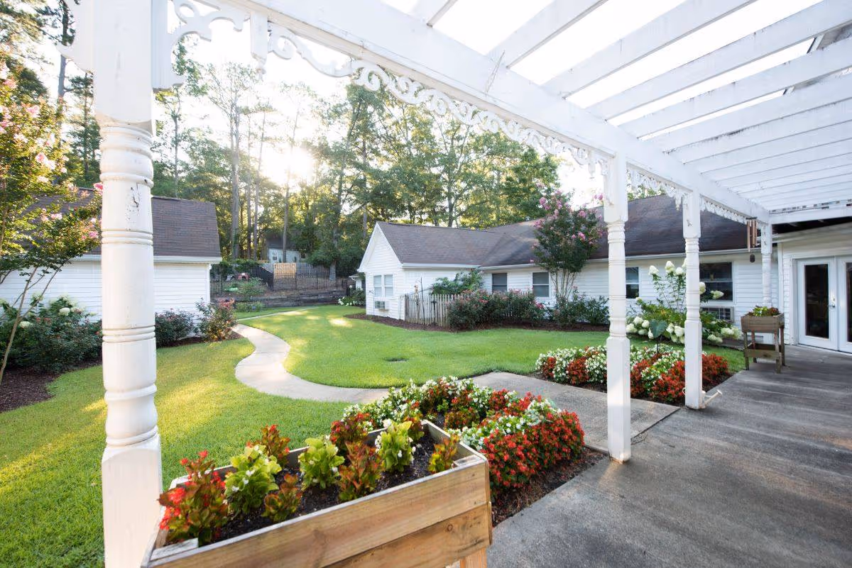Sunlit courtyard with a pergola-covered walkway, flowerbeds, and small white single-story buildings.