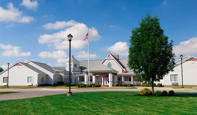 Front exterior view of a single-story senior living facility building with white siding, a gray roof, and a central entrance with columns. There is a flagpole with an American flag in front, a large tree on the right, and well-maintained green grass and shrubs surrounding the building under a partly cloudy blue sky.