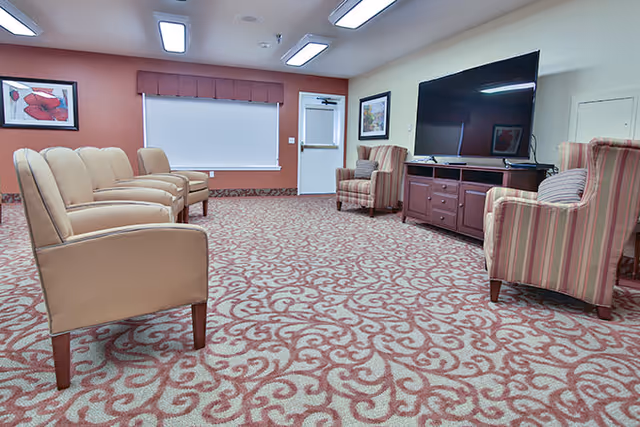 Community sitting room with rows of upholstered chairs facing a large flat-screen TV on a console, patterned carpet and framed artwork.
