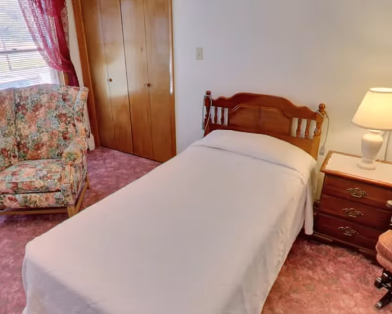 A small bedroom with a single wooden bed covered with a white sheet, a wooden nightstand with a lamp, a floral upholstered armchair, a window with red curtains, and a wooden closet with double doors. The floor is covered with pink carpet.