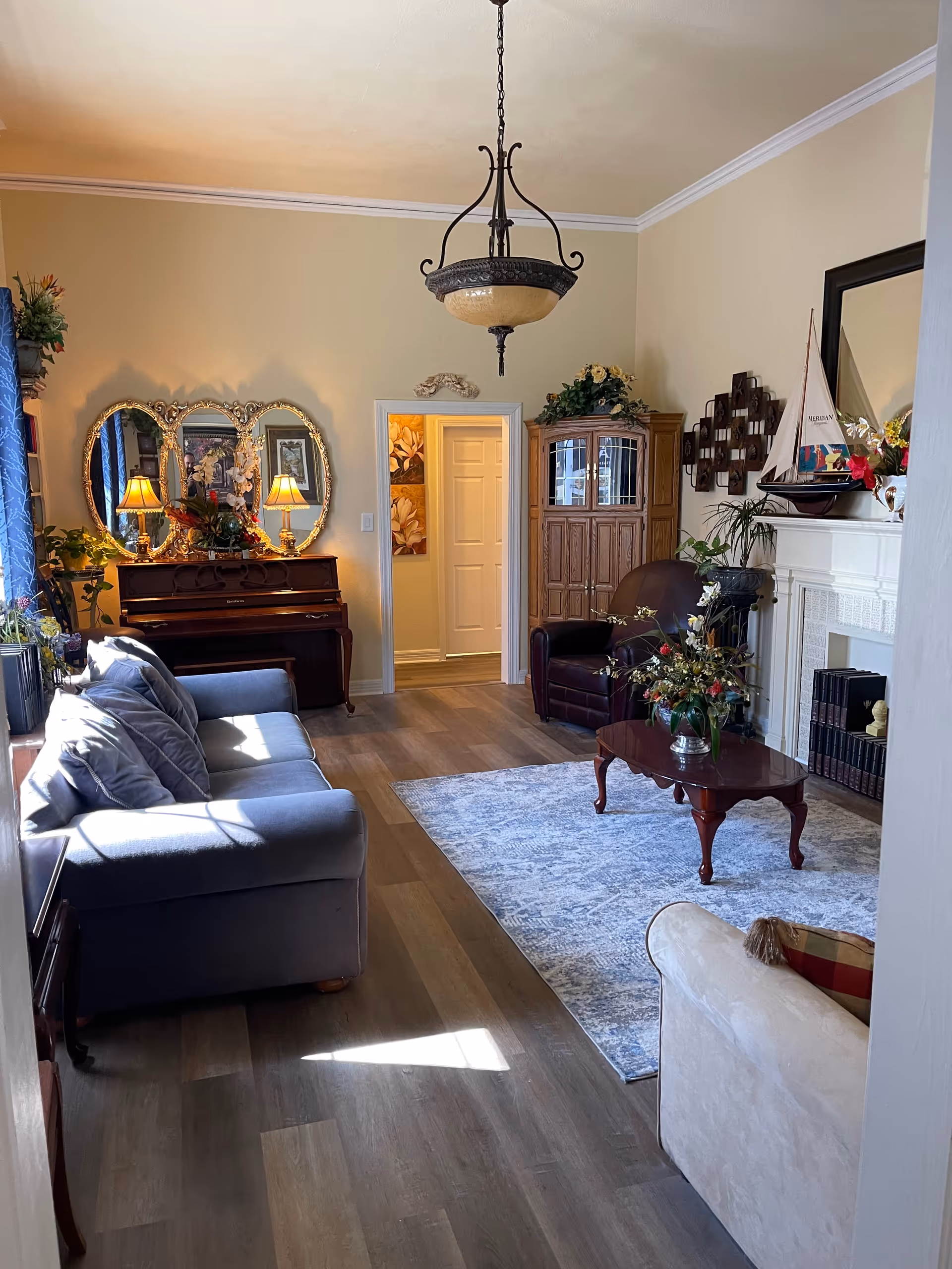 A bright, traditionally furnished living room featuring a sofa and armchairs, a piano, coffee table on a rug, decorative mirrors, and a chandelier.
