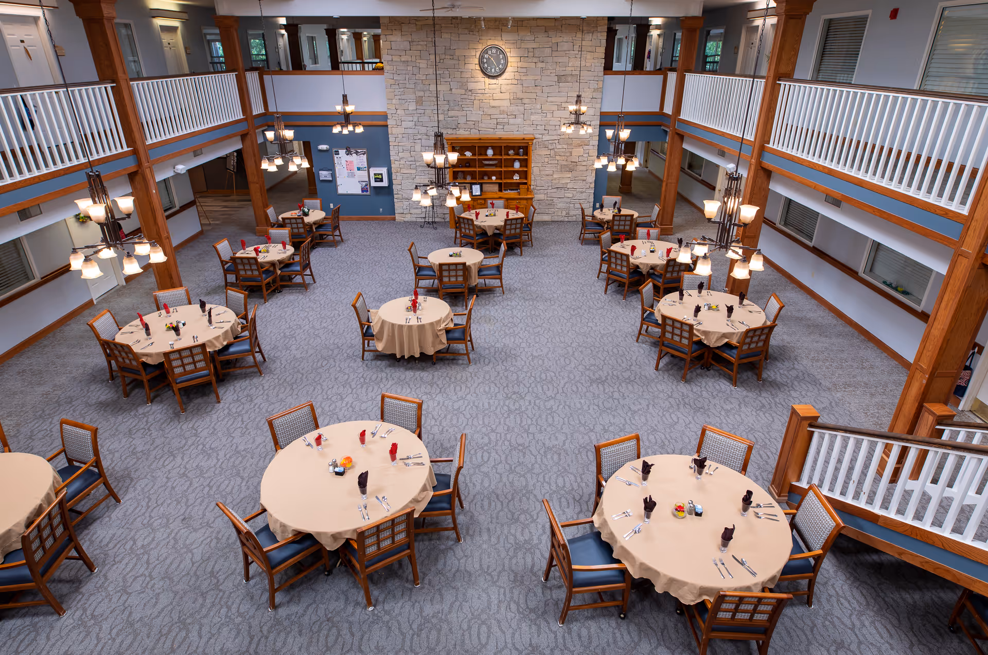 Spacious dining room with multiple round tables covered with beige tablecloths, each set with napkins, utensils, and condiments. The room features a high ceiling with hanging chandeliers, a stone wall with a clock, wooden pillars, and a second-floor balcony overlooking the area.