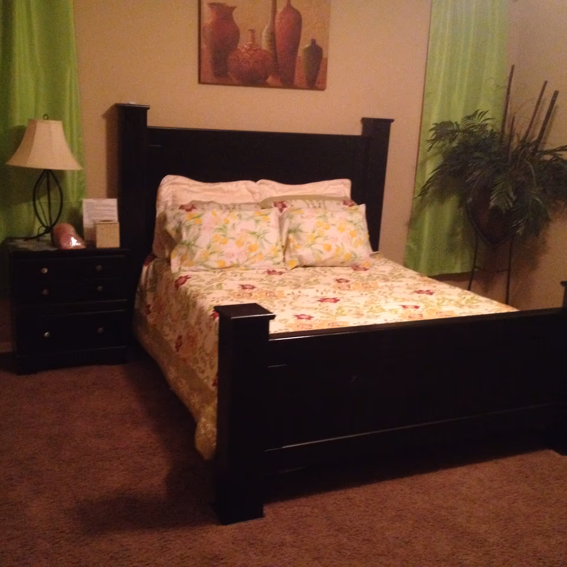 A bedroom featuring a dark wood bed with floral bedding, a nightstand with a lamp, green curtains and a potted plant.