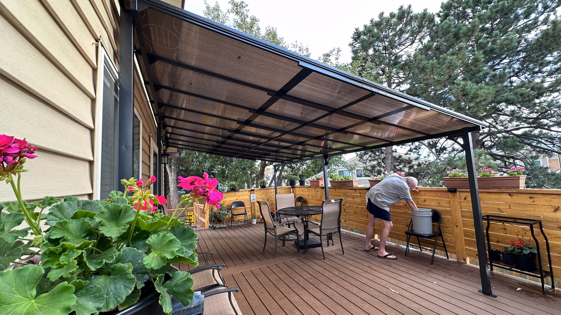 A covered outdoor patio area with wooden flooring and a transparent roof. There are several chairs and a round table on the patio. A man is bending over near a chair with a bucket on it. The patio is surrounded by a wooden fence with flower pots placed on top. Trees and neighboring houses are visible in the background.