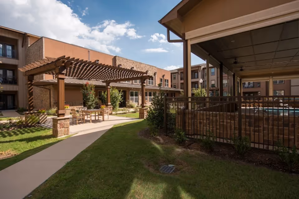 Outdoor courtyard area at a senior living facility with a wooden pergola covering a seating area with tables and chairs, surrounded by green grass and plants. The building with multiple windows and balconies is visible in the background under a partly cloudy sky.