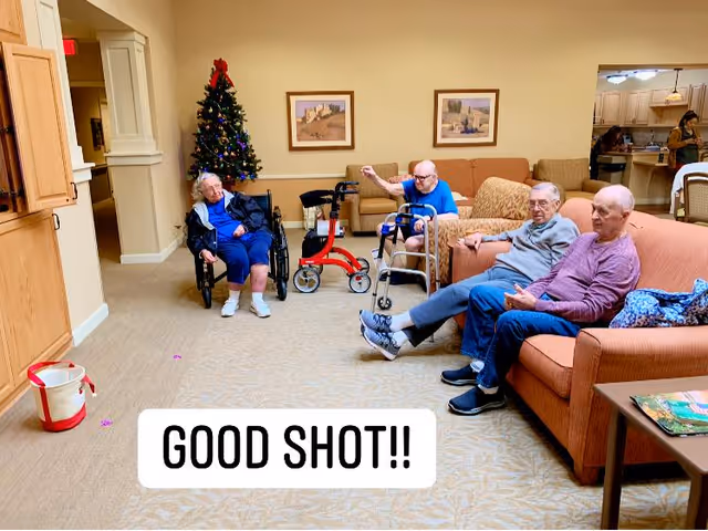 A common area in a senior living facility with four elderly men seated; one in a wheelchair near a decorated Christmas tree, two sitting on a couch, and one sitting on a chair. The room has beige walls, framed artwork, and a carpeted floor. In the background, a woman is visible in a kitchen area.