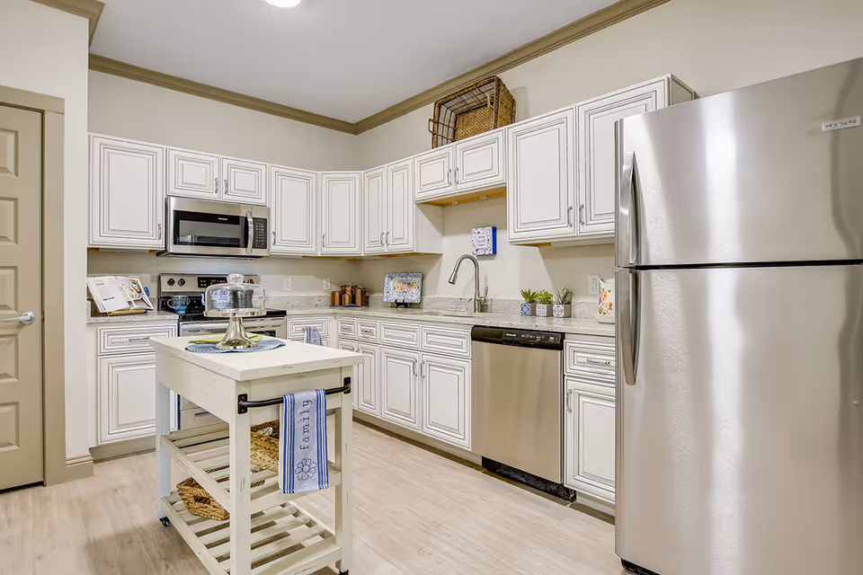 A modern kitchen with white cabinetry, stainless steel appliances including a refrigerator, microwave, stove, and dishwasher. There is a small white kitchen island with a towel hanging on the side and a cake stand on top. The kitchen has light wood flooring and beige walls with crown molding.
