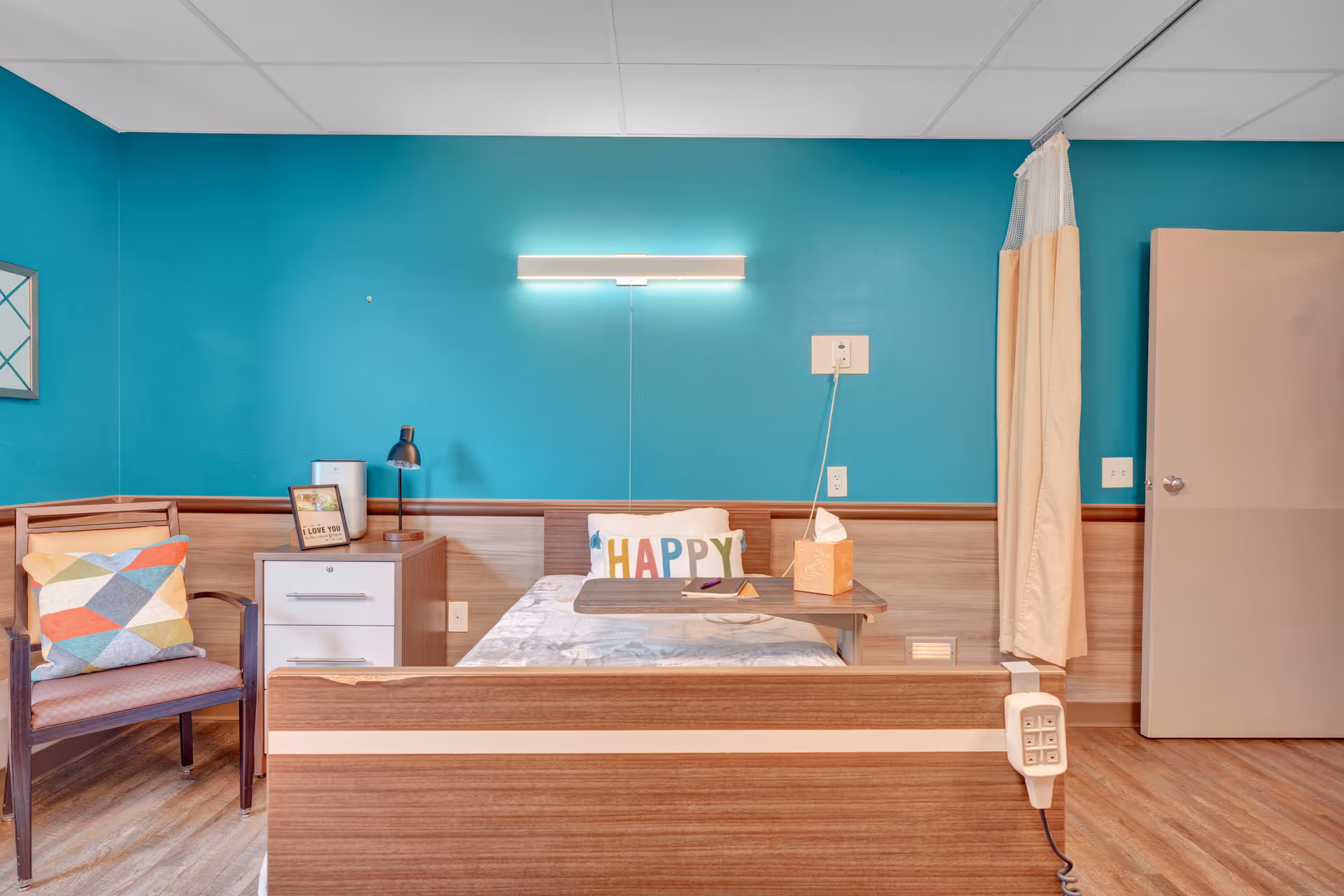 A neatly arranged nursing facility room with a single bed featuring a 'HAPPY' pillow and a tray table with tissues and a remote. The room has teal walls, a wooden chair with a colorful cushion, a small white nightstand with a lamp and a framed picture, and a beige privacy curtain partially drawn to the side. The floor is wooden, and there is a closed beige door on the right.
