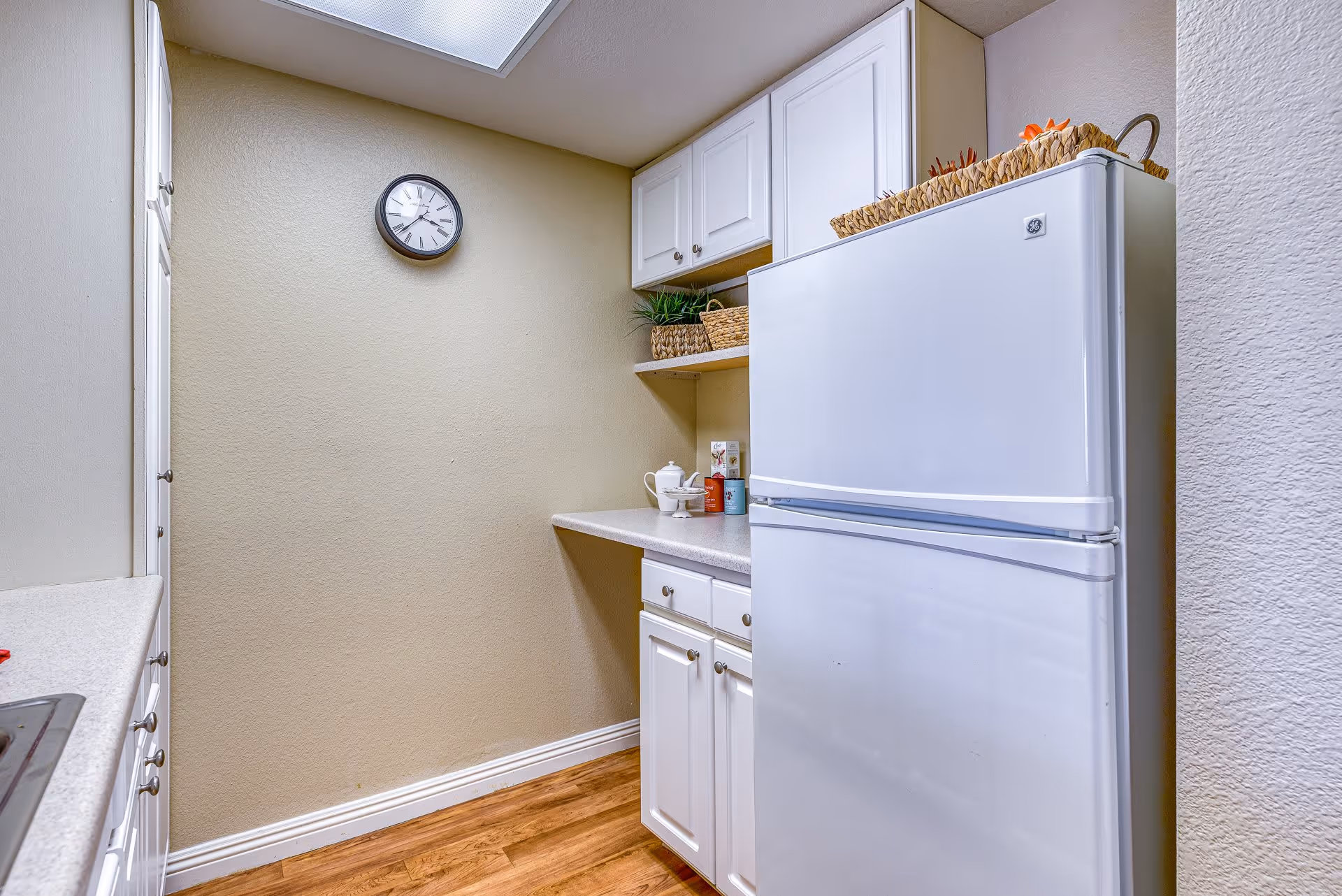 A small kitchen area with white cabinets, a white refrigerator, and a countertop. There are baskets and small decorative items on the shelves and countertop. A clock is mounted on the beige wall, and the floor is wooden.