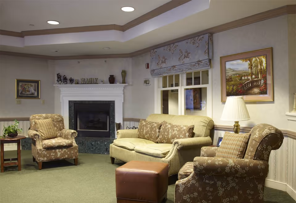 A cozy living room area with a beige sofa and two patterned armchairs, a brown leather ottoman, a side table with a plant, a white fireplace with decorative items on the mantel, and framed artwork on the walls.