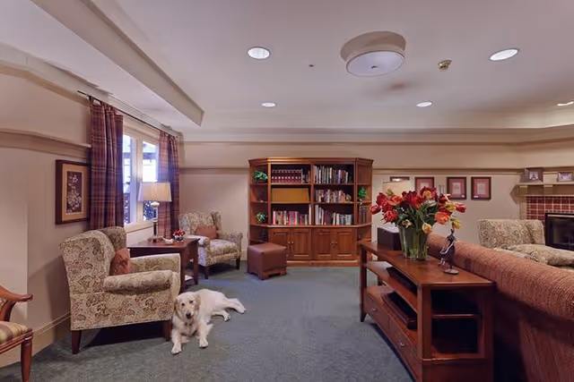 A cozy living room with patterned armchairs, a wooden bookshelf filled with books, a wooden coffee table with a vase of red flowers, and a golden retriever dog lying on the carpeted floor. The room has soft lighting, a window with plaid curtains, and framed pictures on the walls.
