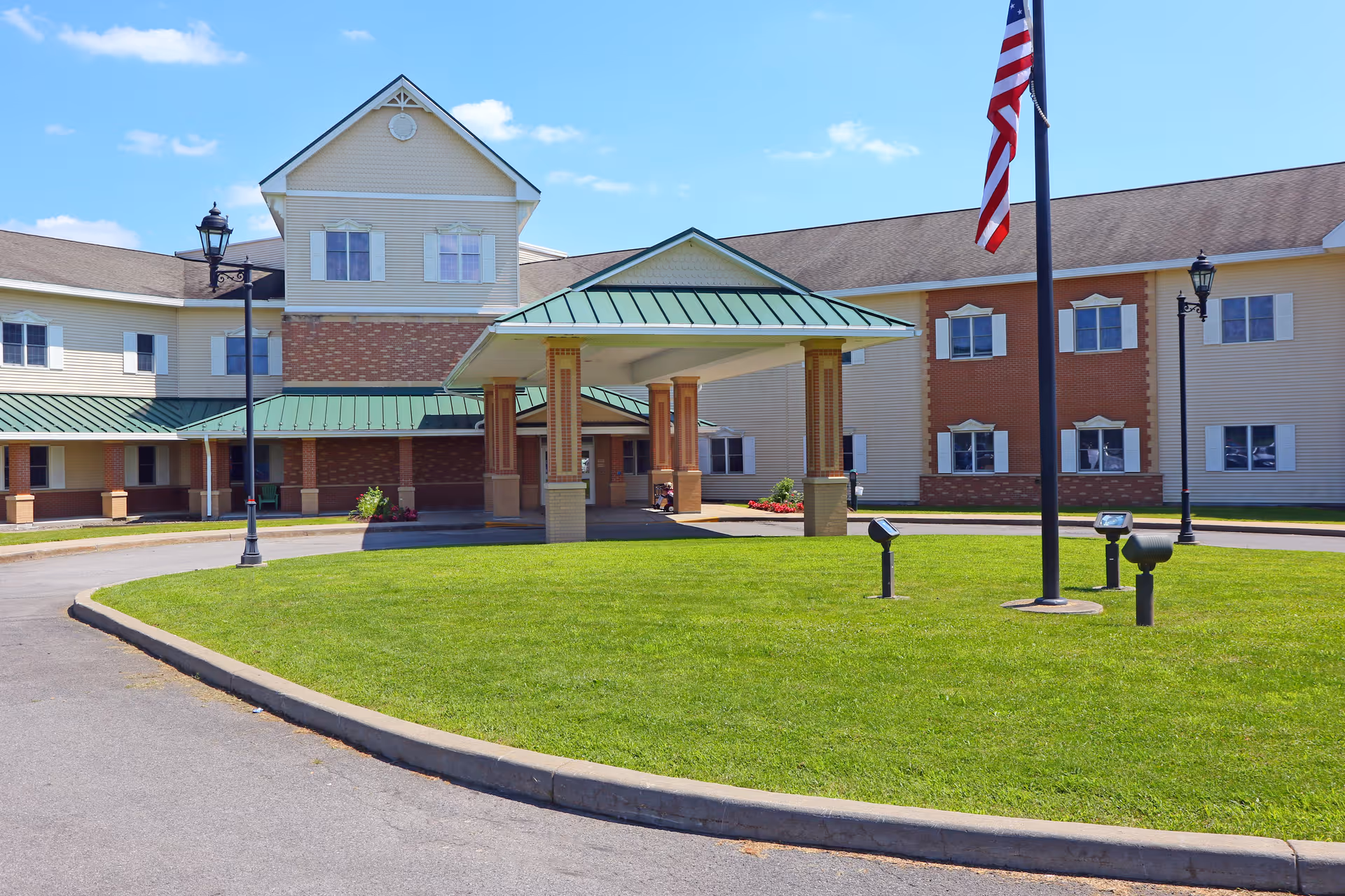 Front exterior view of Cooperstown Center, a multi-story building with beige and brick facade, green metal roof over the entrance, a circular driveway, and an American flag on a flagpole in the foreground.