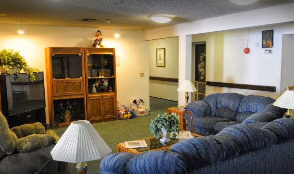 A cozy living room area with blue upholstered sofas arranged around a wooden coffee table with a plant and newspapers. There are two table lamps with white shades, a wooden cabinet with glass doors displaying decorative items, a TV in the corner, and a green armchair. The room has carpeted floors and white walls with some framed artwork.