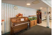 Interior view of a hallway area in a senior living facility featuring a wooden upright piano with a floral arrangement on top, a fish tank on a wooden stand, a bookshelf with decorative items, and floral wallpaper with a clock above the piano.