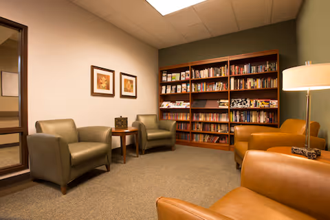 A cozy reading room with two green armchairs and two brown leather armchairs arranged around a small wooden table. A tall wooden bookshelf filled with books and magazines is against the wall. The room has beige carpet, neutral-colored walls, two framed pictures, and a floor lamp providing warm lighting.