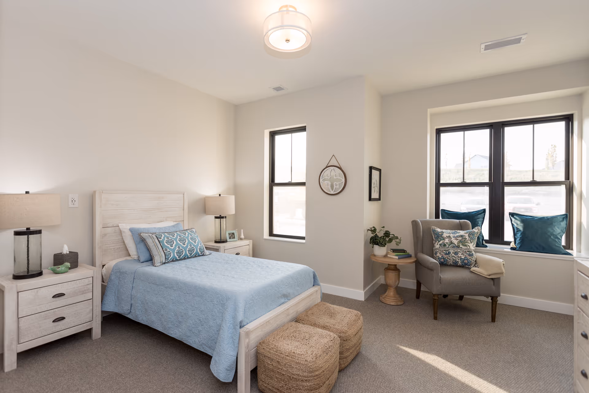A bright and cozy bedroom with a light wood bed frame and matching nightstands on either side. The bed is made with a light blue quilt and decorative pillows. Two woven ottomans are placed at the foot of the bed. There is a gray armchair with patterned and solid blue pillows near two large windows letting in natural light. A small round wooden side table with a plant and books is next to the chair. The walls are painted light beige, and the room has a soft carpeted floor.