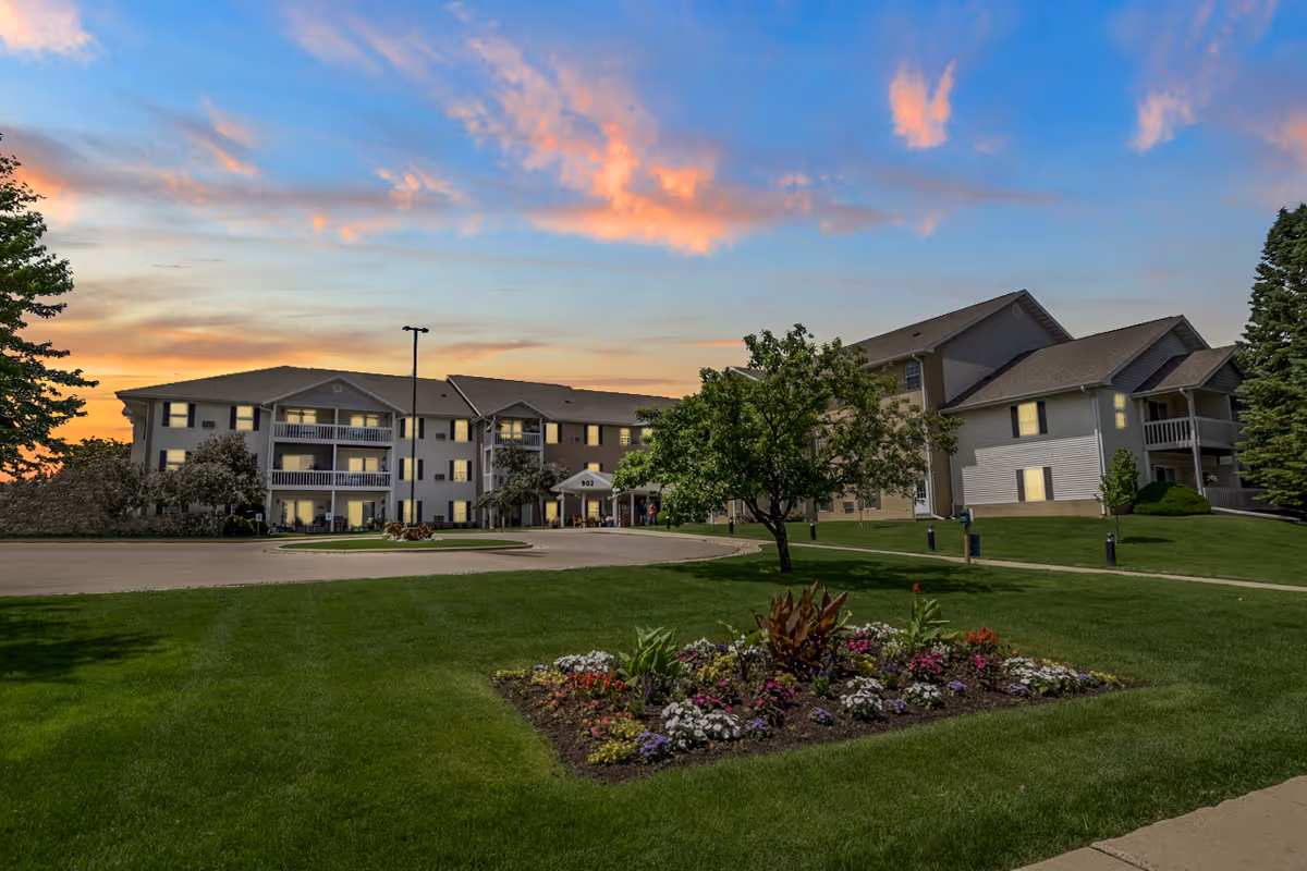 Exterior view of a senior housing and apartment community building at sunset with a well-maintained lawn, a flower bed, trees, and a driveway leading to the entrance.