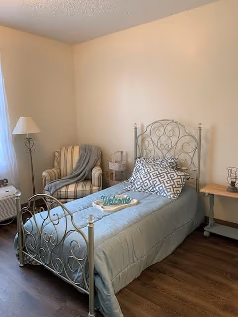 A cozy senior living bedroom with a single metal bed frame, light blue bedding, and a decorative pillow. A 'Welcome' sign is placed on the bed. Next to the bed is a small wooden side table with a decorative item. In the corner, there is a striped armchair with a gray throw blanket draped over it and a floor lamp beside it. The room has wooden flooring and light-colored walls.