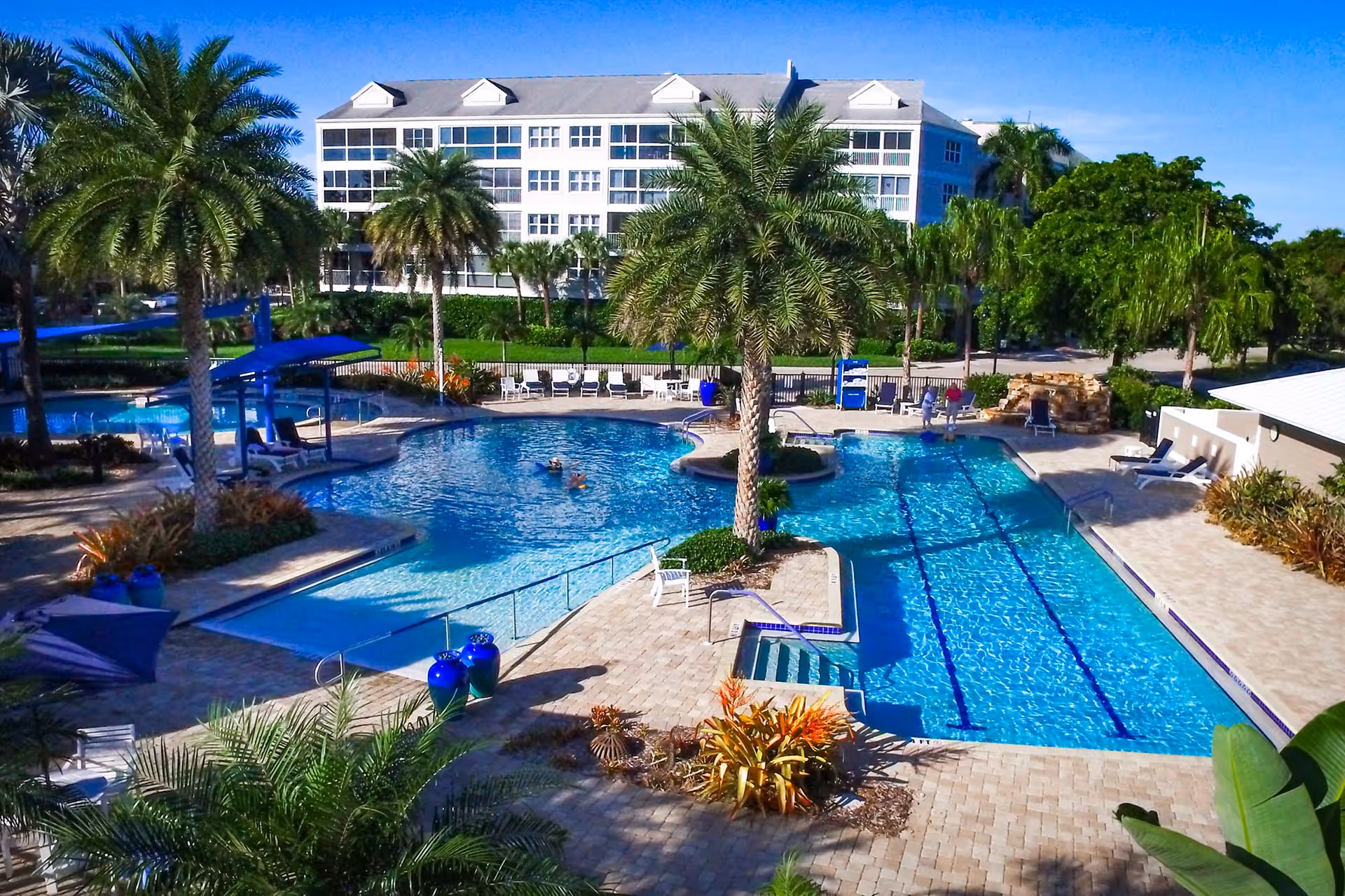 Outdoor swimming pool area with two connected pools, surrounded by palm trees and lounge chairs. A multi-story white building is visible in the background under a clear blue sky.