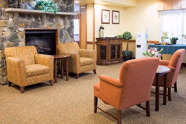 A cozy living room area with four upholstered armchairs arranged around two small wooden tables. There is a stone fireplace with a wooden mantle decorated with a green plant. In the background, there is a wooden cabinet with a coffee dispenser on top, framed botanical prints on the wall, a water cooler, and a table with a green tablecloth and floral arrangements near a window with sheer curtains.