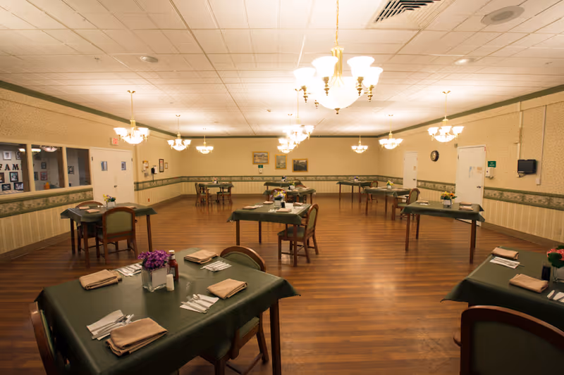 A spacious dining room with several tables covered in green tablecloths, each set with napkins, silverware, and small flower arrangements. The room has wooden flooring, beige wallpaper with a green border, and multiple chandeliers hanging from the ceiling providing warm lighting.