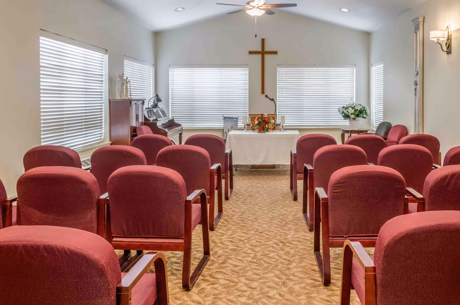 Small chapel-style interior with rows of red upholstered chairs facing a table and a cross at the front.