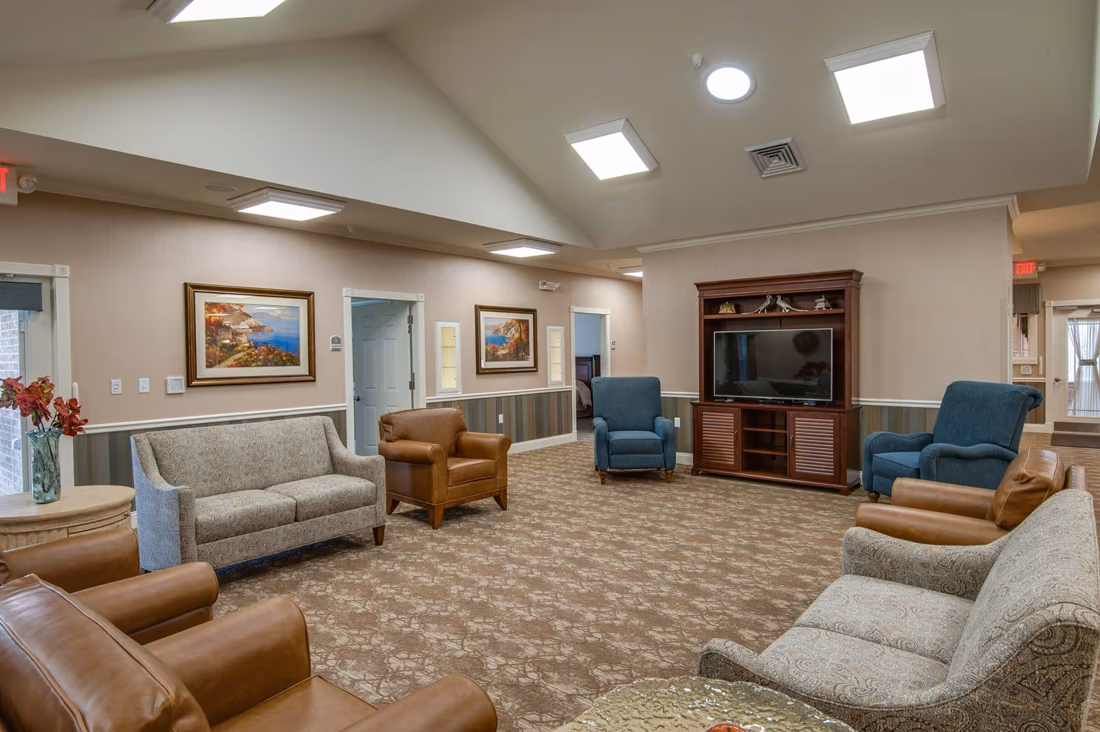 A spacious senior living facility common area with multiple armchairs and sofas arranged around a wooden entertainment center with a flat-screen TV. The room has beige walls with framed landscape paintings, patterned carpet, and ceiling lights including skylights. There is a small round table with a vase of flowers near the entrance.
