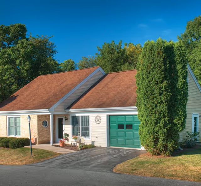 Single-story house with a red roof, white siding, a green garage door, and a small front porch with potted plants. There is a tall evergreen tree on the right side and other trees in the background under a clear blue sky.