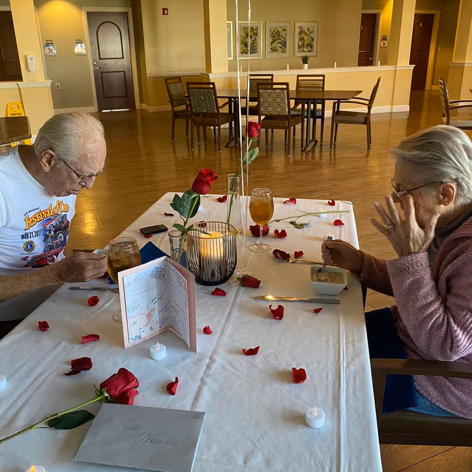 Two elderly people seated at a table decorated with rose petals, candles, and cards in a communal dining area.