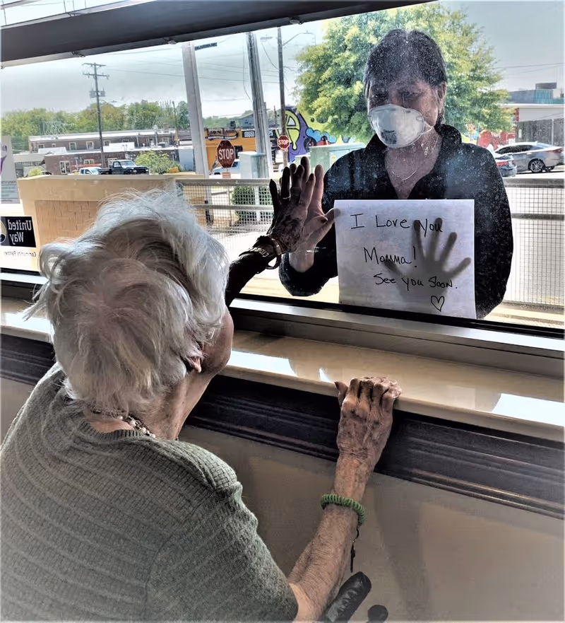 An elderly woman inside a facility presses her hand against a glass window, touching hands with a woman outside who is wearing a face mask and holding a handwritten sign that says 'I Love You Momma! See you soon.'