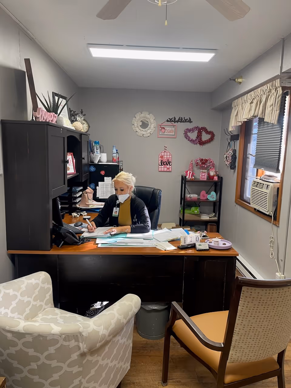 A woman wearing a mask is seated at a desk in a small office, writing on papers. The office has a dark wooden desk with various documents and office supplies on it, a black hutch with decorations including a pink 'love' sign, and a small shelving unit with pink and heart-themed decor. There are two chairs in front of the desk, a window with an air conditioning unit, and a ceiling fan above.