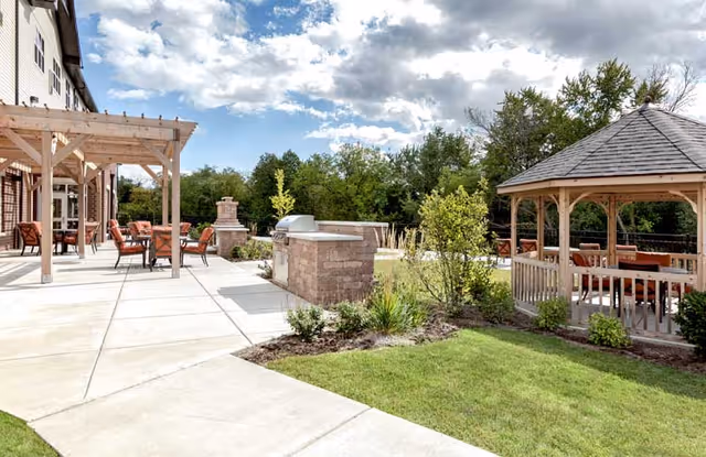 Outdoor patio area with pergolas, a gazebo, seating, and a built-in grill beside a landscaped lawn under a partly cloudy sky.