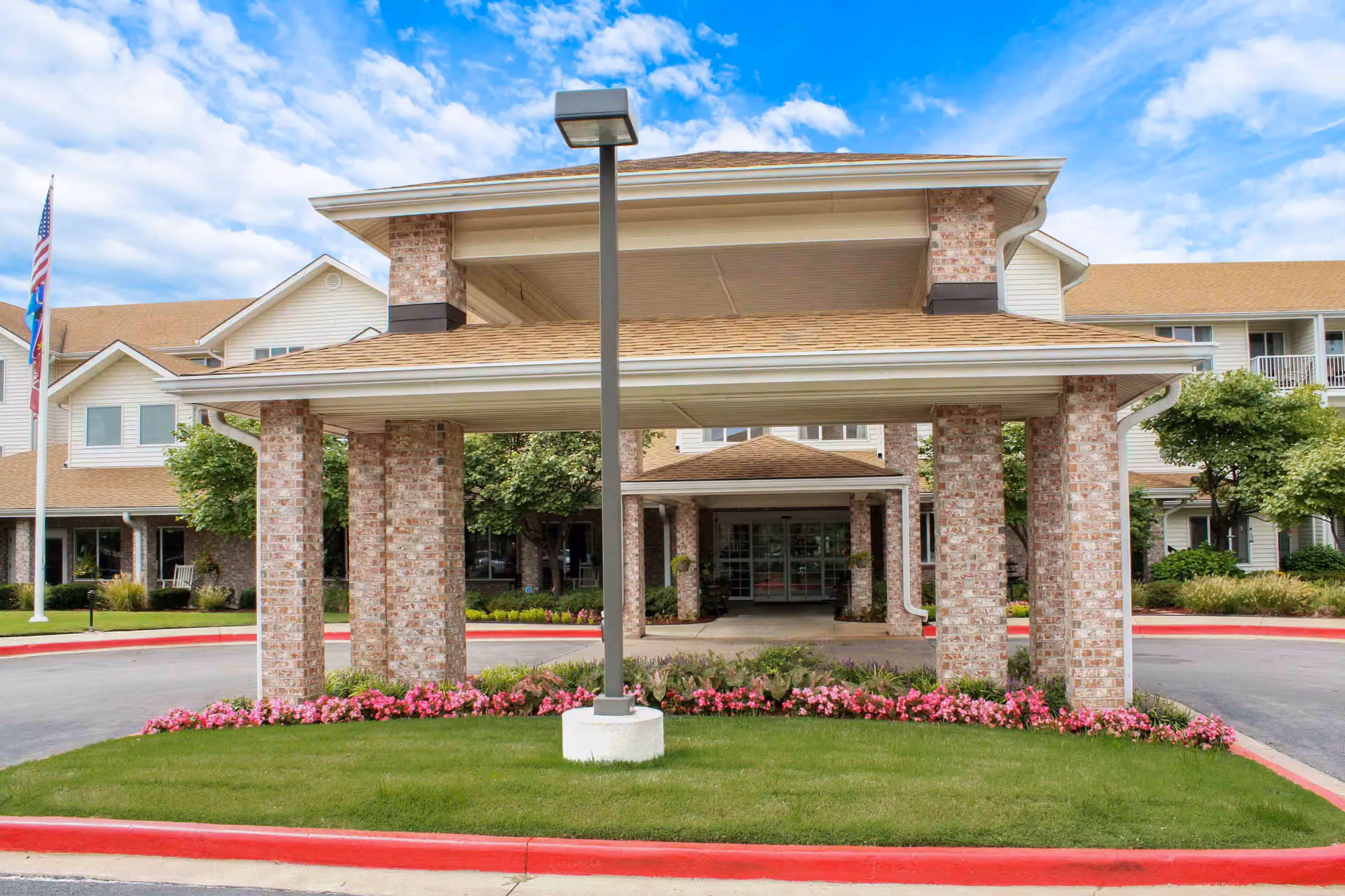 Front entrance of Asher Point Independent Living of Bartlesville featuring a covered driveway with brick pillars, a well-maintained lawn with pink flowers, and an American flag on a flagpole to the left. The building has beige siding and a tan roof under a partly cloudy blue sky.