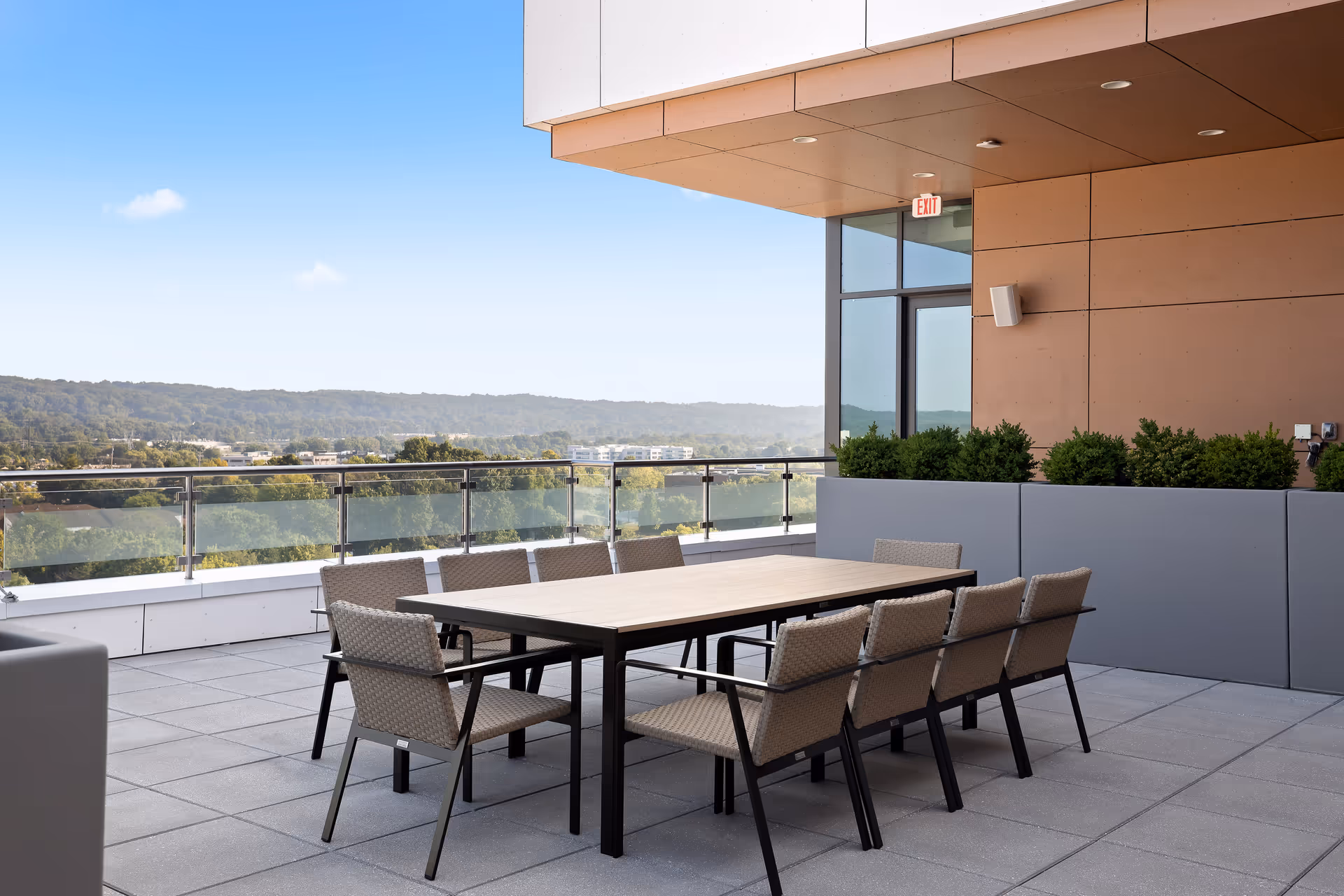 Outdoor patio area with a rectangular table surrounded by eight woven chairs. The patio has a glass railing with a view of trees and hills in the distance under a clear blue sky. There are large planters with green shrubs along the wall of the building.