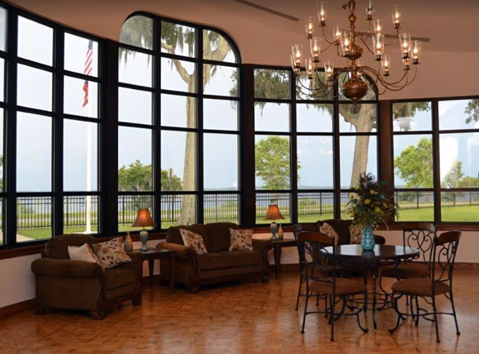 Sunlit communal living area with large arched windows overlooking trees and water, furnished with sofas, a round table and chairs, lamps, and a chandelier.