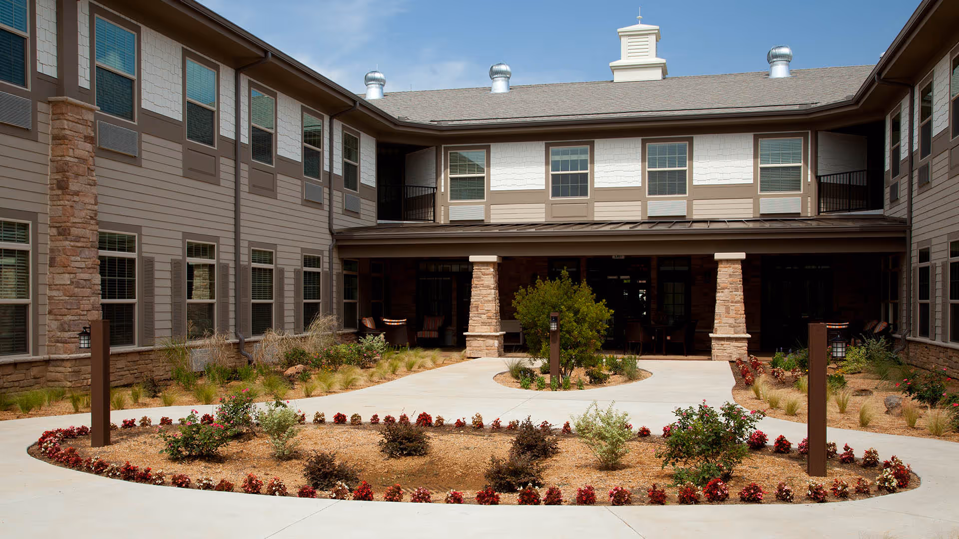 Landscaped courtyard and entrance of a two-story senior living building with a circular flower bed and paved walkways.
