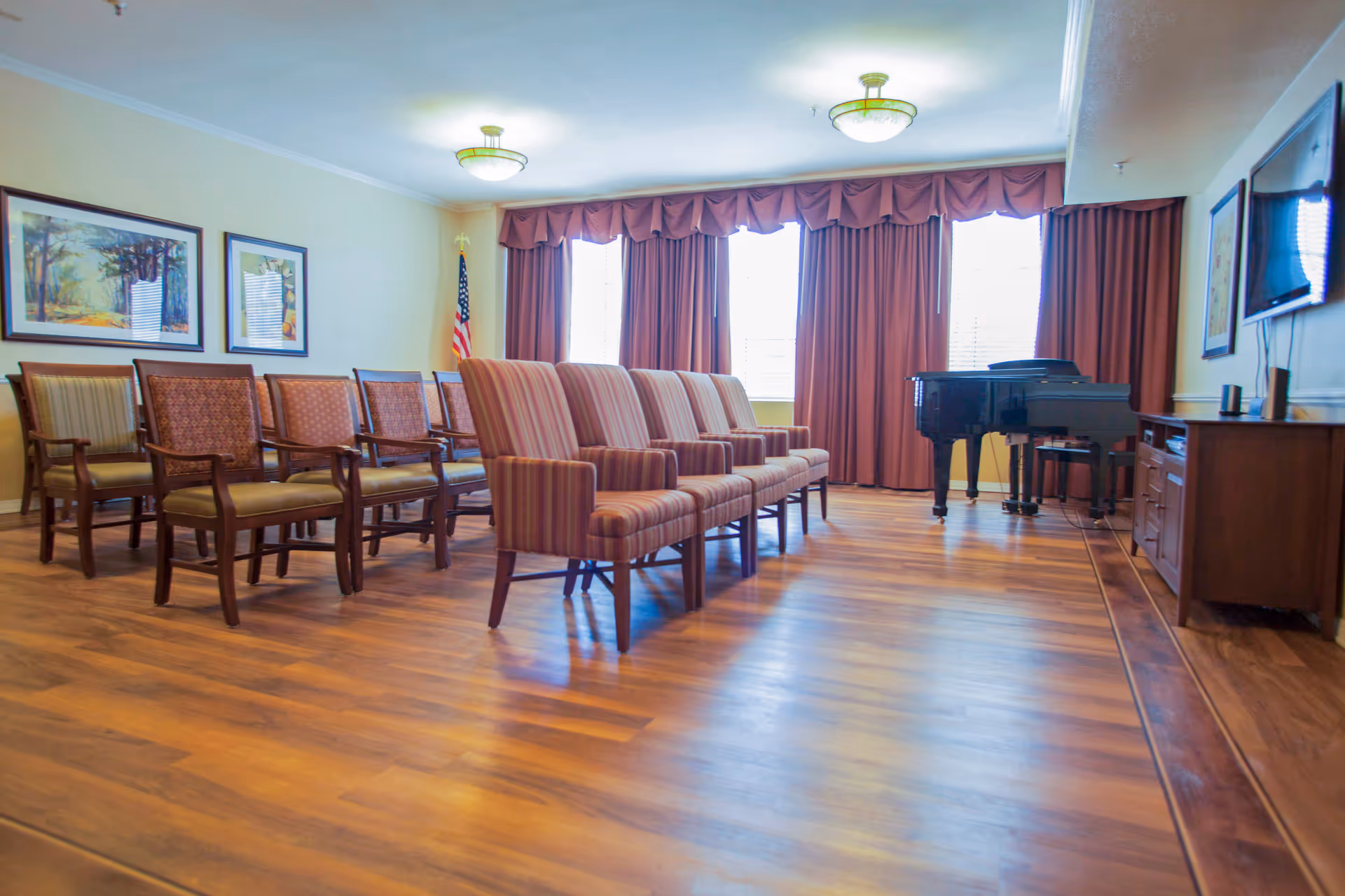 A spacious common room with rows of upholstered chairs facing a grand piano and wall-mounted TV, hardwood floors and curtained windows.