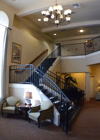 Interior view of a senior living facility featuring a carpeted staircase with black metal railings leading to an upper floor. Two upholstered chairs and a small round table with a lamp are positioned near a large window with curtains. A chandelier hangs from the ceiling, and framed artwork is displayed on the walls.
