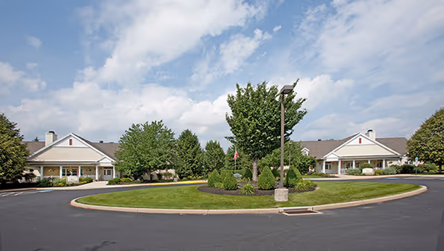 Front exterior view of a senior living facility with a circular driveway, landscaped greenery, trees, and a lamppost under a partly cloudy sky.