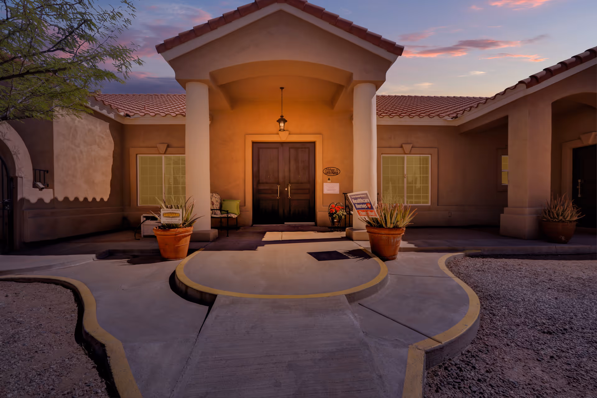 Entrance of Lovin Manor facility at dusk with a covered porch supported by two white columns, double wooden doors, potted plants, and signs near the doorway. The building has a tiled roof and beige stucco walls.