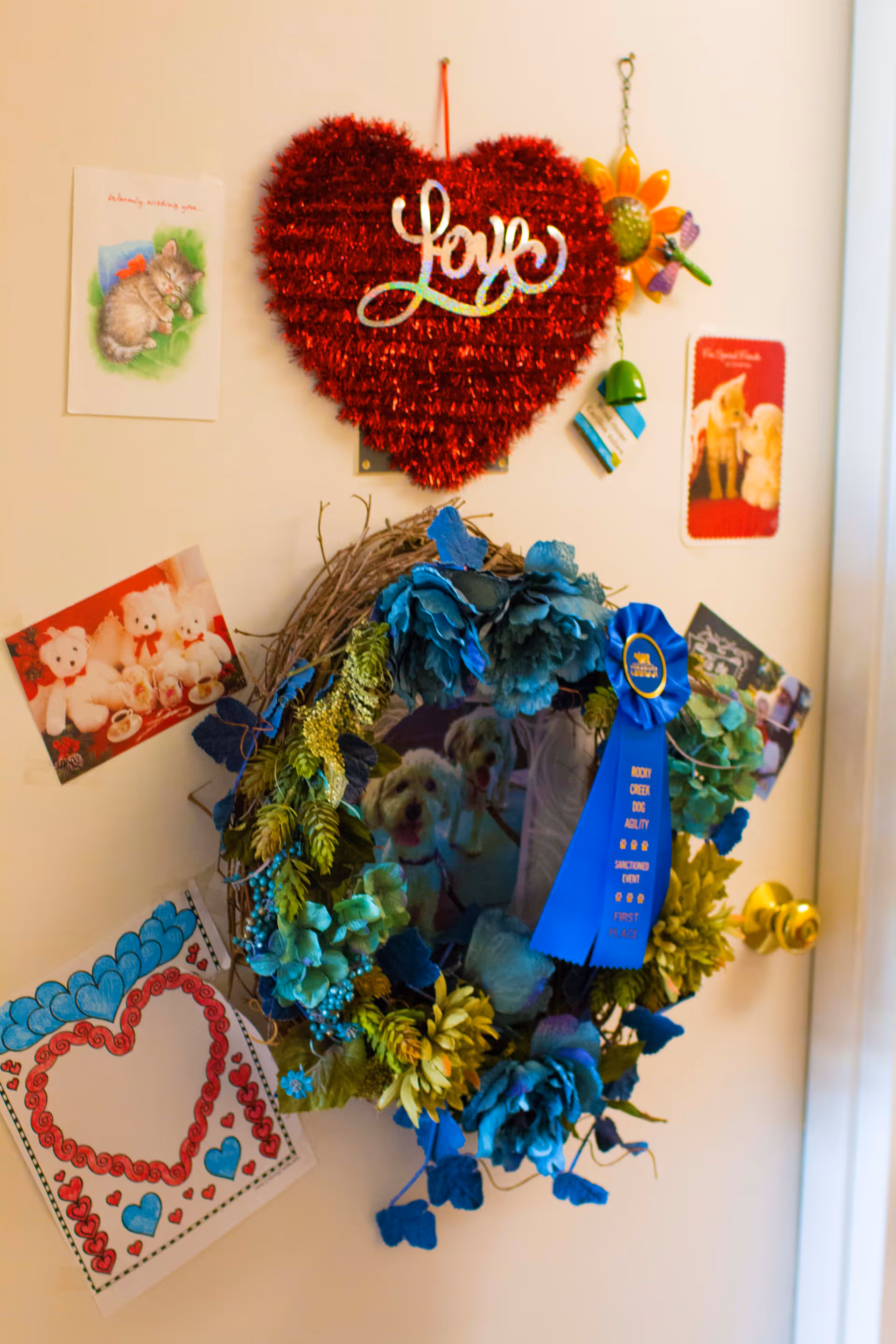 A decorated door with a red tinsel heart, a floral wreath adorned with a blue award ribbon, and several greeting cards and photos.