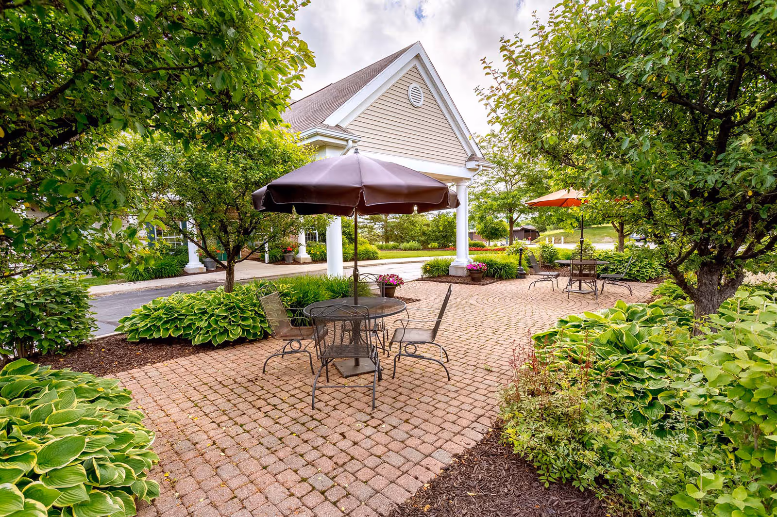 Brick-paved outdoor patio with metal tables, chairs and umbrellas surrounded by greenery in front of a building entrance.
