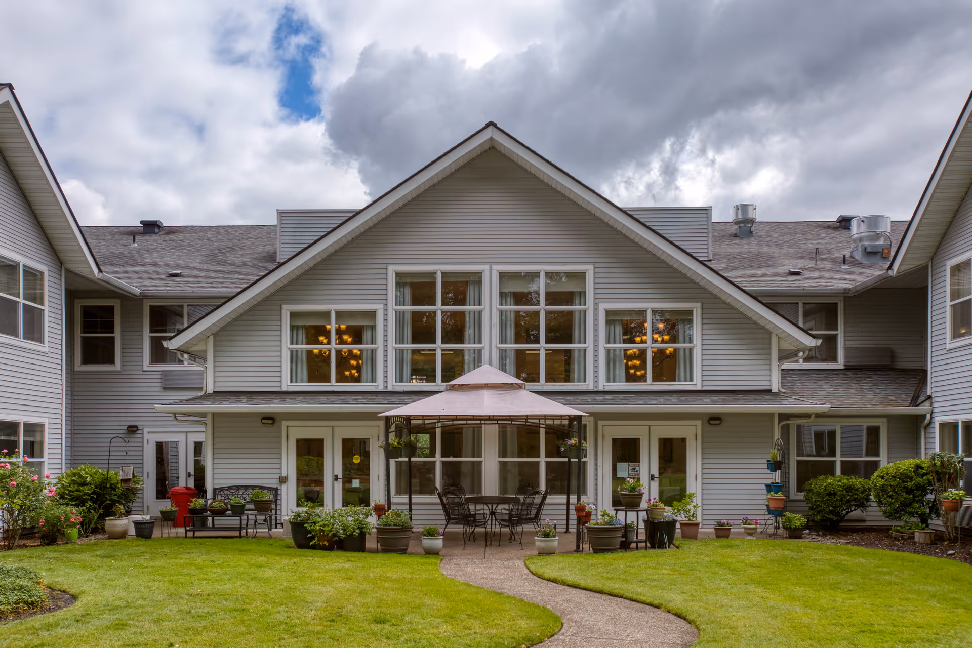 Exterior view of a two-story assisted living facility with gray siding, large windows, and a covered patio area with outdoor seating and potted plants. The building is surrounded by a well-maintained lawn and shrubbery under a cloudy sky.