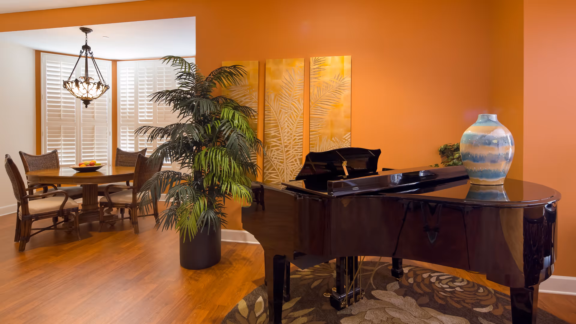 A cozy interior space featuring a glossy black grand piano with a decorative vase on top, positioned on a patterned rug. Behind the piano is an orange wall with three vertical golden leaf art panels. To the left, there is a dining area with a round wooden table, four wicker chairs, a bowl of fruit on the table, and a hanging chandelier above. A large potted plant is placed near the piano, and white plantation shutters cover the windows in the background.