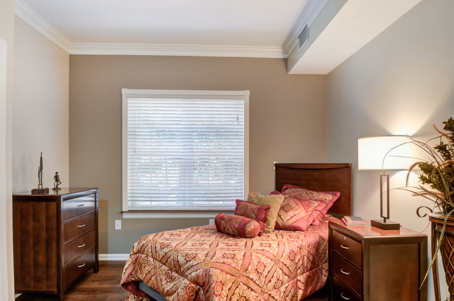A cozy single bedroom with a patterned bedspread, wooden headboard, nightstand and dresser next to a window with blinds.