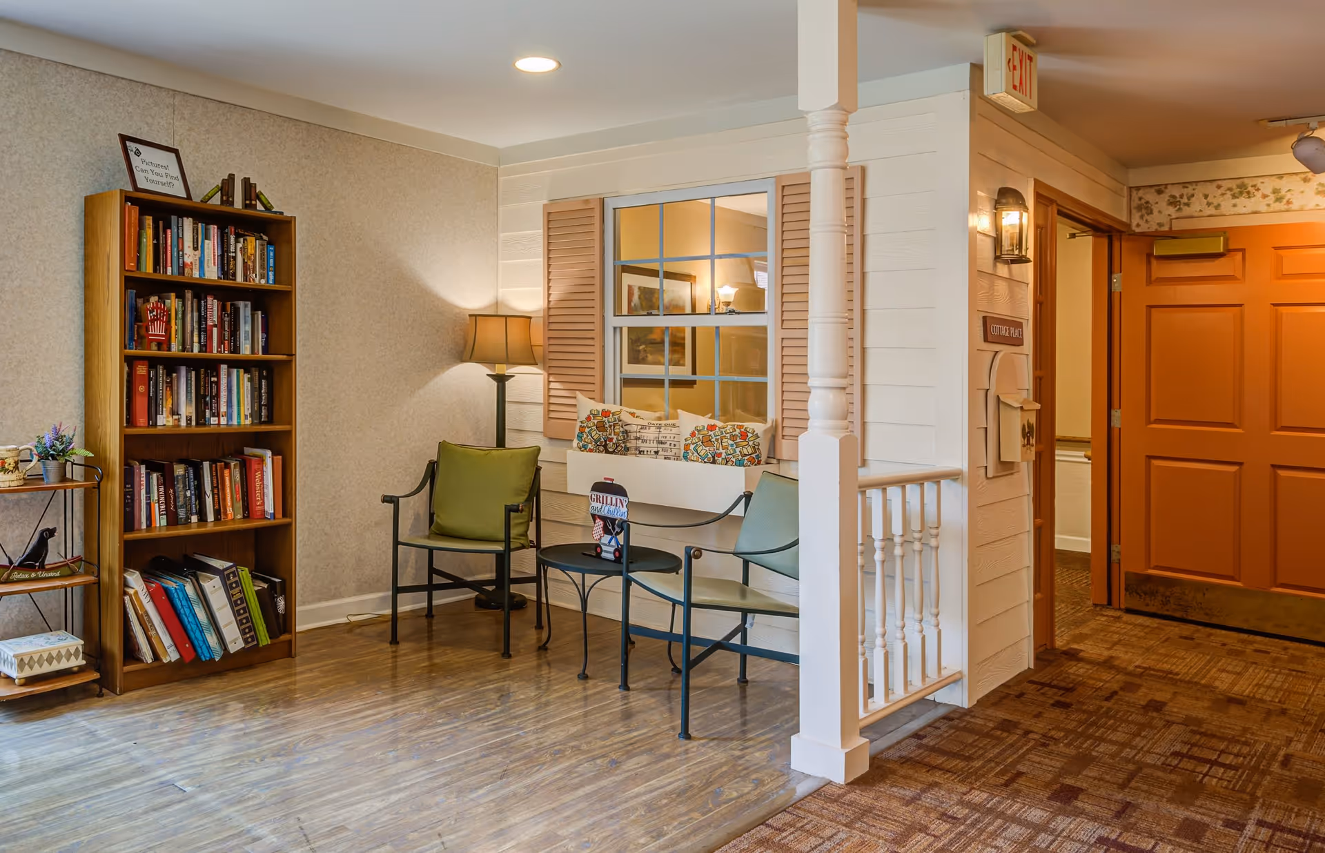 A cozy interior corner of a senior living facility featuring a wooden bookshelf filled with books, a small metal side table with a plant and decorative items, two green cushioned chairs with a small round table between them, and a white bench with colorful pillows. The area has wood flooring and a carpeted hallway leading to an orange door. A window with wooden shutters and a lamp are visible on the wall behind the seating area.